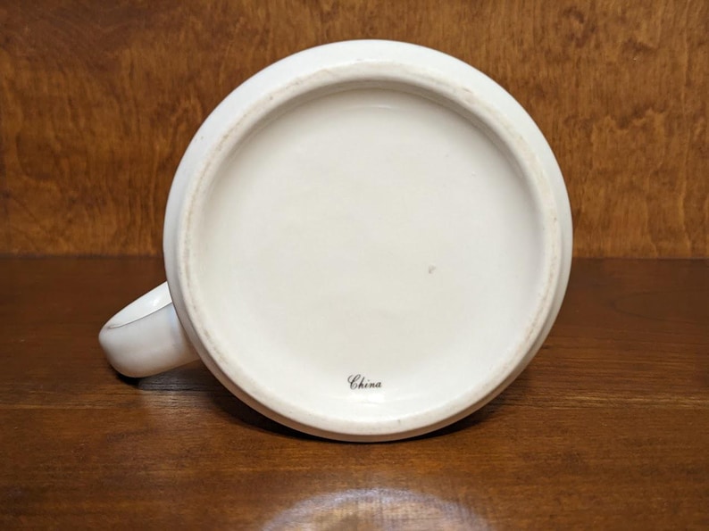 Puede incluir: Una taza de cer&aacute;mica blanca, vista desde abajo, con una base redondeada y un asa peque&ntilde;a. La palabra "China" est&aacute; impresa en escritura negra cerca del centro. La taza est&aacute; sobre una superficie de madera oscura.