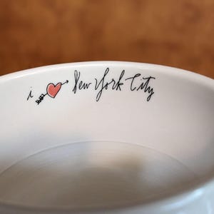 May include: A white ceramic bowl with the phrase "i ❤️ New York City" in black script. A red heart pierced by a black arrow is between the "i" and "New". The bowl has a smooth, glossy finish.