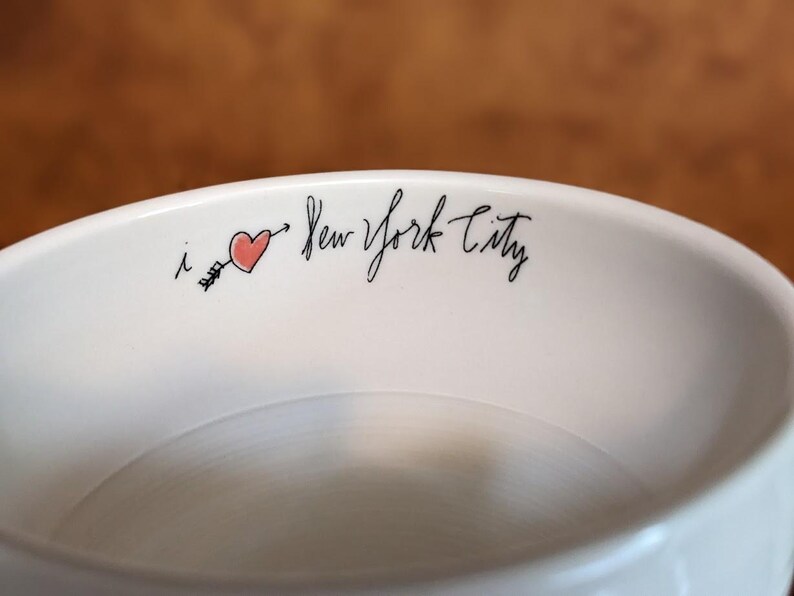 May include: A white ceramic bowl with the phrase "i ❤️ New York City" in black script. A red heart pierced by a black arrow is between the "i" and "New". The bowl has a smooth, glossy finish.