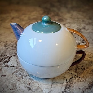 Könnte beinhalten: Eine weiße Teekanne aus Keramik mit einem türkisfarbenen Deckel und einem goldenen Henkel. Die Teekanne hat einen runden Korpus und einen violetten Ausguss.