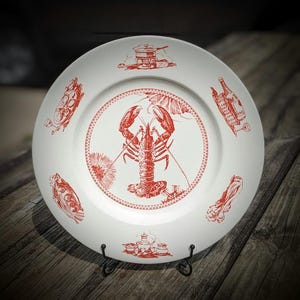 Peut inclure: Assiette en céramique blanche avec une illustration de homard rouge au centre, entourée d'autres dessins rouges liés à la nourriture. L'assiette a une bordure décorative et est présentée sur un support en métal noir. L'assiette est sur une surface en bois.