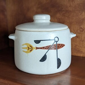 Peut inclure: Pot en céramique crème avec couvercle et poignées latérales. Il présente un motif fantaisiste de fourchette, de cuillère et de cuillères à mesurer en jaune, marron et noir. Le pot est posé sur une surface en bois.