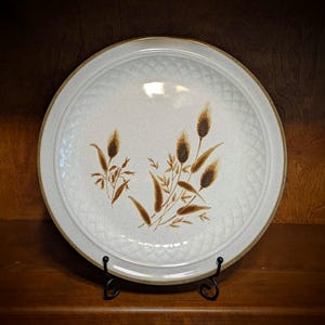 May include: A round, off-white ceramic plate with a brown floral design. The plate has a textured rim and a brown border. The floral design features brown wheat stalks and leaves. The plate is displayed on a black metal stand.