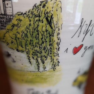 May include: White ceramic mug with a hand-drawn illustration of a New York City scene, including a building and a weeping willow tree. The mug has the text "NYC I love you" with a red heart.