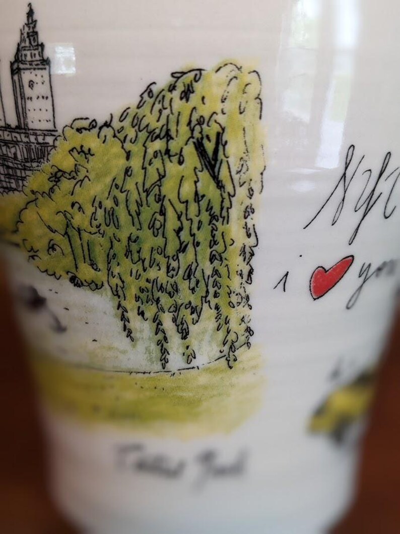 May include: White ceramic mug with a hand-drawn illustration of a New York City scene, including a building and a weeping willow tree. The mug has the text "NYC I love you" with a red heart.