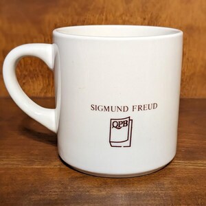 Puede incluir: Una taza de cer&aacute;mica blanca con un asa curva. La taza presenta el nombre "SIGMUND FREUD" en letras granates, junto con un peque&ntilde;o logotipo. La taza est&aacute; sobre un fondo de madera.