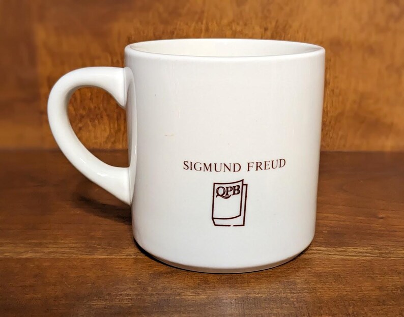 Puede incluir: Una taza de cer&aacute;mica blanca con un asa curva. La taza presenta el nombre "SIGMUND FREUD" en letras granates, junto con un peque&ntilde;o logotipo. La taza est&aacute; sobre un fondo de madera.