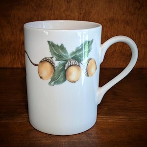 May include: A white ceramic mug featuring a painted illustration of three brown acorns with textured caps attached to a green leafy branch. The mug has a smooth cylindrical body and a curved handle on the right side, showcasing a clean design.