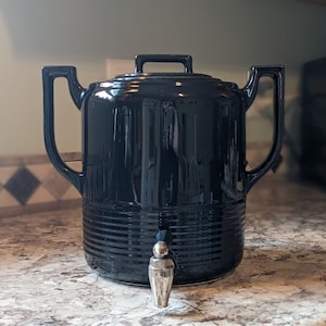 May include: A black ceramic beverage dispenser with a spigot. The dispenser has a rounded body with a ribbed design and two handles.