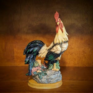 May include: A rooster figurine featuring a cream-colored head and neck, a bright red comb and wattle, and dark blue-green tail feathers. The bird stands on a textured gray rock base with small green foliage accents, mounted on a round wooden plinth.
