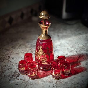 May include: A ruby red glass decanter with a gold-toned metal stopper and neck, featuring gold-painted figures in period attire. The set includes five matching small ruby red glass tumblers, each decorated with gold-painted scenes and gold rims.