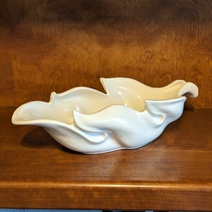 May include: A white ceramic bowl with a unique, organic shape resembling flowing leaves. The bowl is smooth and has a glossy finish.