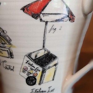 May include: Cream-colored ceramic mug featuring a hand-drawn illustration of an Italian ice cart with a red and white umbrella. The cart has the words "Italian Ices" and "Italian Ice Cart" written on it. The mug has a curved handle.