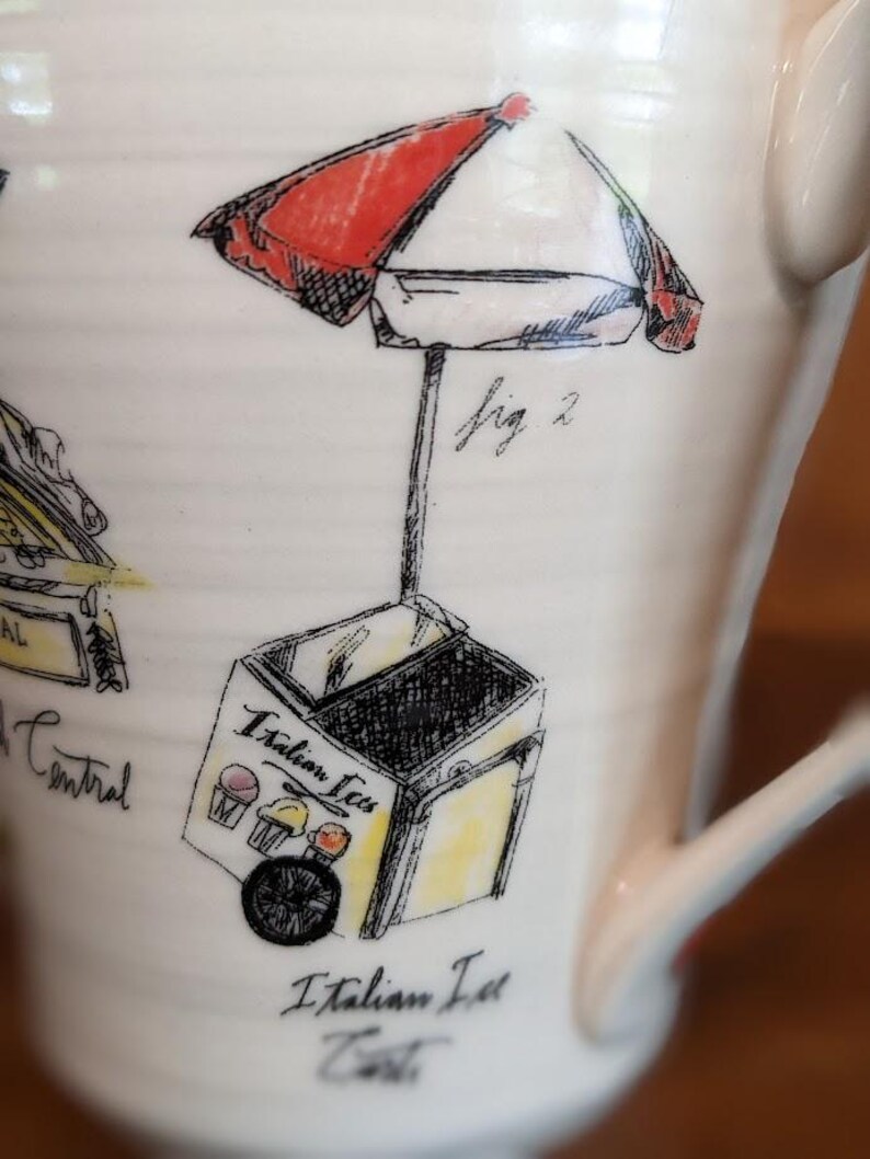 May include: Cream-colored ceramic mug featuring a hand-drawn illustration of an Italian ice cart with a red and white umbrella. The cart has the words "Italian Ices" and "Italian Ice Cart" written on it. The mug has a curved handle.