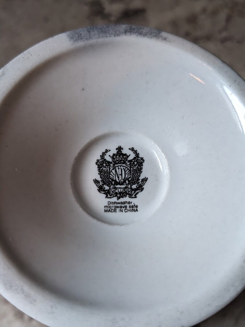 May include: White ceramic mug with a black and white crest featuring a crown and the letters "NH" with the text "Exclusive" underneath. The bottom of the mug reads "Dishwasher, microwave safe MADE IN CHINA."