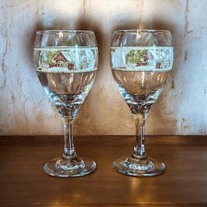 May include: Two clear wine glasses with a winter scene design around the top. The design features a red barn, trees, and snow. The glasses have a long stem and a wide base, perfect for serving wine.