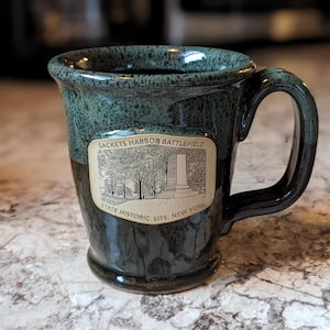 Puede incluir: Una taza de cerámica con un esmalte moteado verde oscuro y marrón. La taza presenta un asa negra y una etiqueta rectangular con una ilustración y el texto "Sackets Harbor Battlefield State Historic Site, New York."