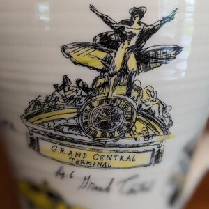 May include: A white ceramic mug featuring a black and yellow illustration of the Grand Central Terminal. The design includes a winged figure, a clock, and the text "GRAND CENTRAL TERMINAL".