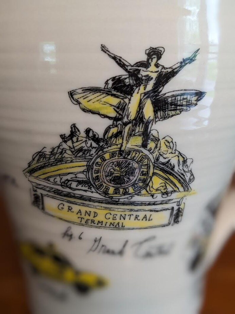 May include: A white ceramic mug featuring a black and yellow illustration of the Grand Central Terminal. The design includes a winged figure, a clock, and the text "GRAND CENTRAL TERMINAL".