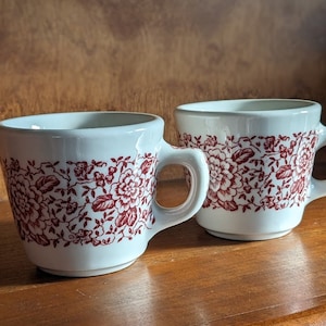Könnte beinhalten: Zwei weiße Keramikbecher mit roten Blumendesigns. Die Becher haben zwei Henkel.
