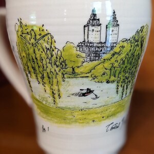 May include: White ceramic mug with a hand-drawn illustration of a park scene. The design features a lake with a person in a rowboat, trees, and a building with two towers. The mug has a curved handle and a wide base.