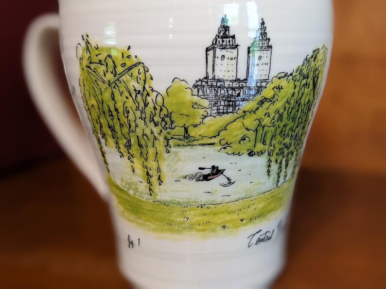 May include: White ceramic mug with a hand-drawn illustration of a park scene. The design features a lake with a person in a rowboat, trees, and a building with two towers. The mug has a curved handle and a wide base.