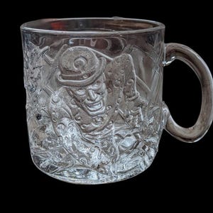 May include: Clear glass mug with a detailed embossed design featuring a character in a hat. The mug has a curved handle and is set against a black background. The design appears to be a vintage or collectible item.