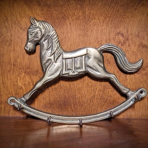 May include: A silver-colored metal rocking horse figurine. The horse has a detailed mane, saddle, and curved rockers. The figurine is displayed against a wooden background, creating a vintage aesthetic.
