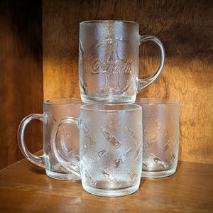 Könnte beinhalten: Vier transparente Coca-Cola-Becher und -Gläser. Die Becher haben Henkel und das Coca-Cola-Logo auf der Vorderseite. Die Gläser haben ein Coca-Cola-Flaschenmuster. Alles steht auf einer Holzoberfläche.