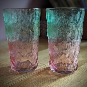 Könnte beinhalten: Zwei hohe, strukturierte Trinkgläser mit Farbverlauf. Die Oberseite jedes Glases ist türkis, geht in einen rosafarbenen Pfirsichton am Boden über. Die Gläser haben eine raue, rindenartige Textur.