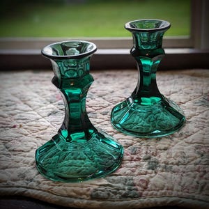 May include: Two emerald green glass candle holders featuring a faceted, multi-sided base and a tapered stem. The glass is translucent with a polished finish, showing a wide circular opening at the top and a stable, flared foot at the bottom.