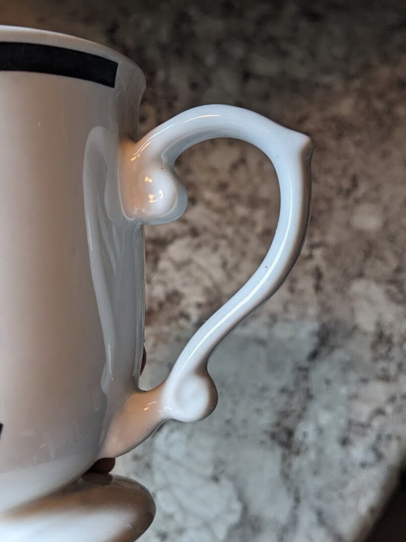 May include: White ceramic mug with a black stripe around the top and a white handle with a unique shape.