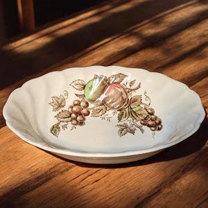 Może przedstawiać: Owalny, kremowy ceramiczny półmisek z falistym brzegiem. Naczynie posiada centralny wzór owoców, w tym winogron, zielonego jabłka i brzoskwini, w odcieniach brązu, zieleni i różu.