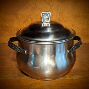 Op de afbeelding: Een roestvrijstalen pot met deksel en twee handvatten. Het deksel heeft een decoratieve handgreep. De handvatten op de pot hebben een decoratief ontwerp. De pot staat op een houten oppervlak.