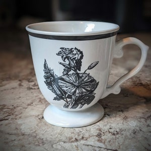 May include: White ceramic mug with a black floral design. The mug has a handle and a pedestal base.
