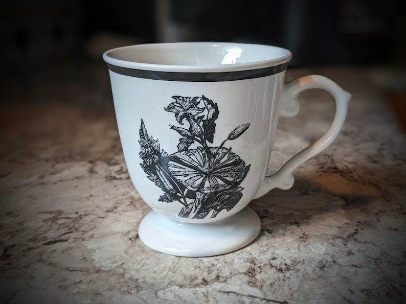 May include: White ceramic mug with a black floral design. The mug has a handle and a pedestal base.
