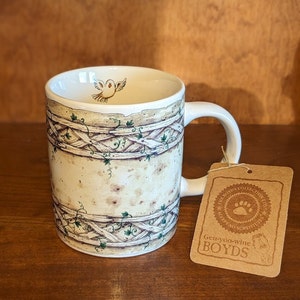 May include: A white ceramic mug with a brown and green design featuring a bird and a vine pattern. The mug has a handle and a brown paper tag with the text "The Boyds Collection - Gen-yoo-wine Boyds - Accept No Substitutes".