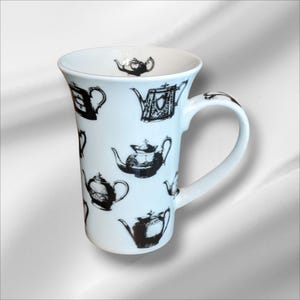 May include: White ceramic mug featuring a repeating pattern of black teapot illustrations. The mug has a curved handle and a wide opening. The teapots vary in style and design, adding visual interest to the mug's surface. A single teapot is also printed inside the mug.