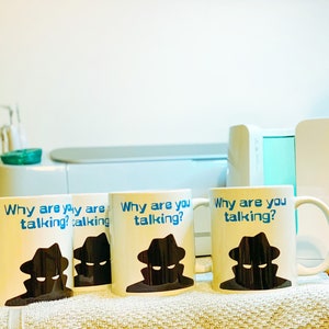 May include: Three white ceramic mugs with blue text that reads "Why are you talking?" and a black silhouette of a person wearing a hat. The mugs are on a white towel.