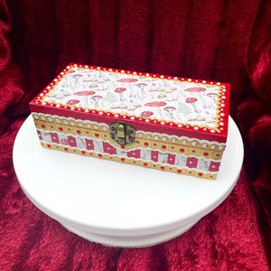 May include: A red wooden box with white and gold trim. The lid is decorated with a pattern of mushrooms and leaves. The box has a small brass latch.