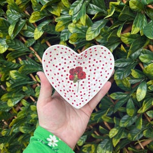 May include: A white heart-shaped trinket dish with red and white polka dots and a red rose design. The dish is being held in a hand wearing a green shirt with white flowers.