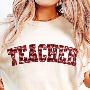 Könnte beinhalten: Cremefarbenes T-Shirt mit dem Wort „TEACHER“ in einem rot glitzernden Design. Das Shirt wird von einer Person mit blonden Haaren und einer blauen Jeans getragen. Die Worte „Digital Download“ stehen in einer lila Box unten links.