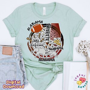 K&ouml;nnte beinhalten: Ein hellgr&uuml;nes T-Shirt mit einem Grafikdesign, das einen Fu&szlig;ball, Erdn&uuml;sse, Elefanten und das Wort "Alabama" mehrmals wiederholt zeigt. Das Design ist in einem Kreis mit einem braunen und roten Rand.
