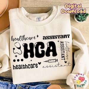 Puede incluir: Una sudadera beige con el texto "healthcare assistant HCA healthcare assistant" en negro. El texto está en un estilo de fuente retro con un estetoscopio, estrellas y un gráfico de corazón.
