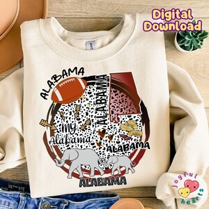 K&ouml;nnte beinhalten: Ein cremefarbener Pullover mit einem Grafikdesign, das den Bundesstaat Alabama, einen Elefanten, einen Football, Erdn&uuml;sse und die Worte "My Alabama" zeigt.