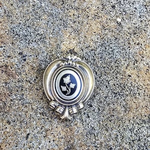May include: A silver pendant with a black and silver floral design. The pendant has an oval shape with a decorative border and a small loop at the top for a chain. The floral design is centered on the pendant.