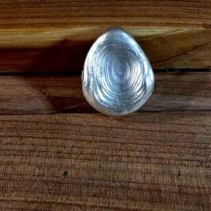 May include: A teardrop-shaped, silver-colored object with a textured, concentric circle design. The object is set against a backdrop of a wooden surface with visible grain patterns. The object appears to be made of metal.