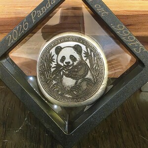 May include: A silver coin featuring a panda bear eating bamboo, displayed in a black, diamond-shaped frame. The frame has the text "2026 Panda" and "2 oz .999 FS" etched on it. The coin is encased in a clear protective cover.