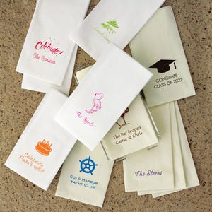 May include: A collection of white paper napkins with various printed designs and text. Designs include a graduation cap, a flamingo, a cake, a ship's wheel, and text that reads "Celebrate!", "The Carsons", "The Byrds", "Congrats Class of 2022", "The Bar is open Carrie & Chris", "Celebrate Paula's 40th!!!", "COLD HARBOR YACHT CLUB", and "The Sterns".