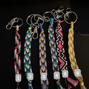May include: Six handmade friendship bracelet keychains with various woven patterns and colors. Each keychain features a metal ring and clasp. The bracelets display a range of colors, including red, blue, yellow, pink, and black, with decorative tassels at the bottom.